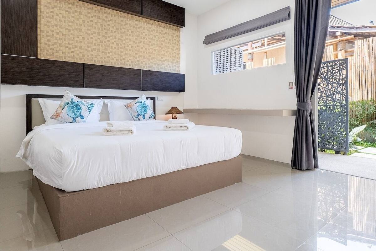 A modern hotel room with a large bed, a headboard with a patterned design, a window with a view, and a balcony with a wooden fence.