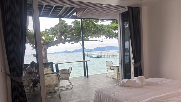 A bedroom with a large bed, a balcony with wicker chairs, and a view of the sea and mountains. - The Strand Resort Samui  (Koh Samui)