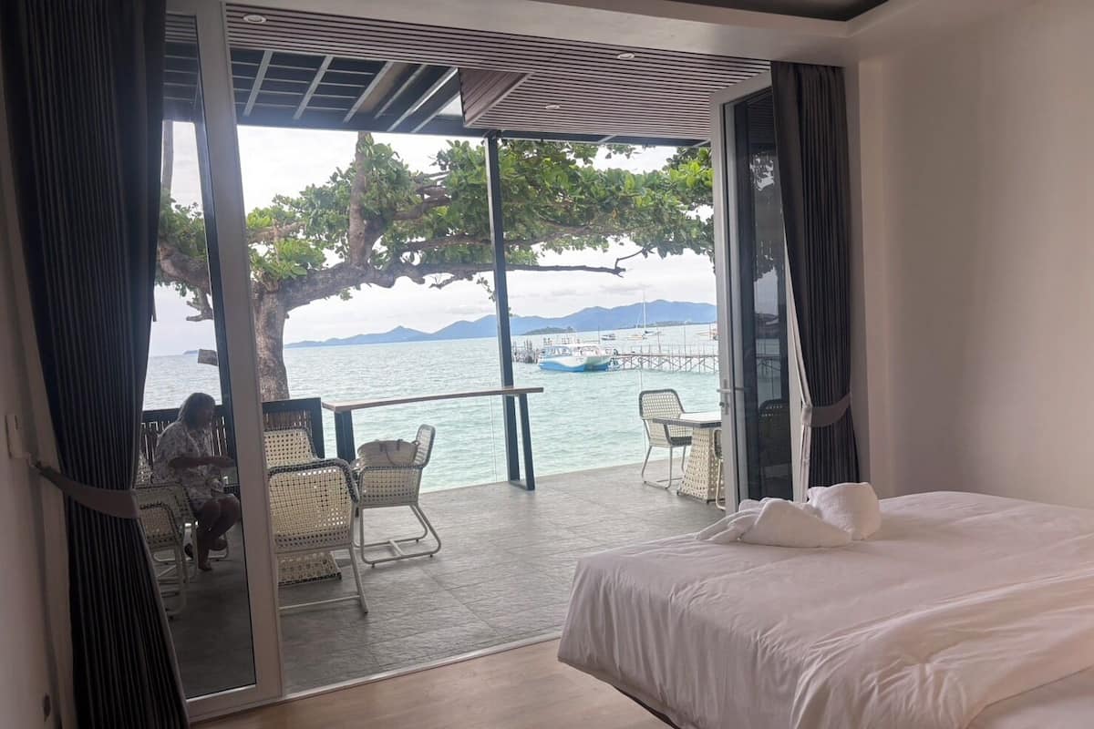 A bedroom with a large bed, a balcony with wicker chairs, and a view of the sea and mountains.
