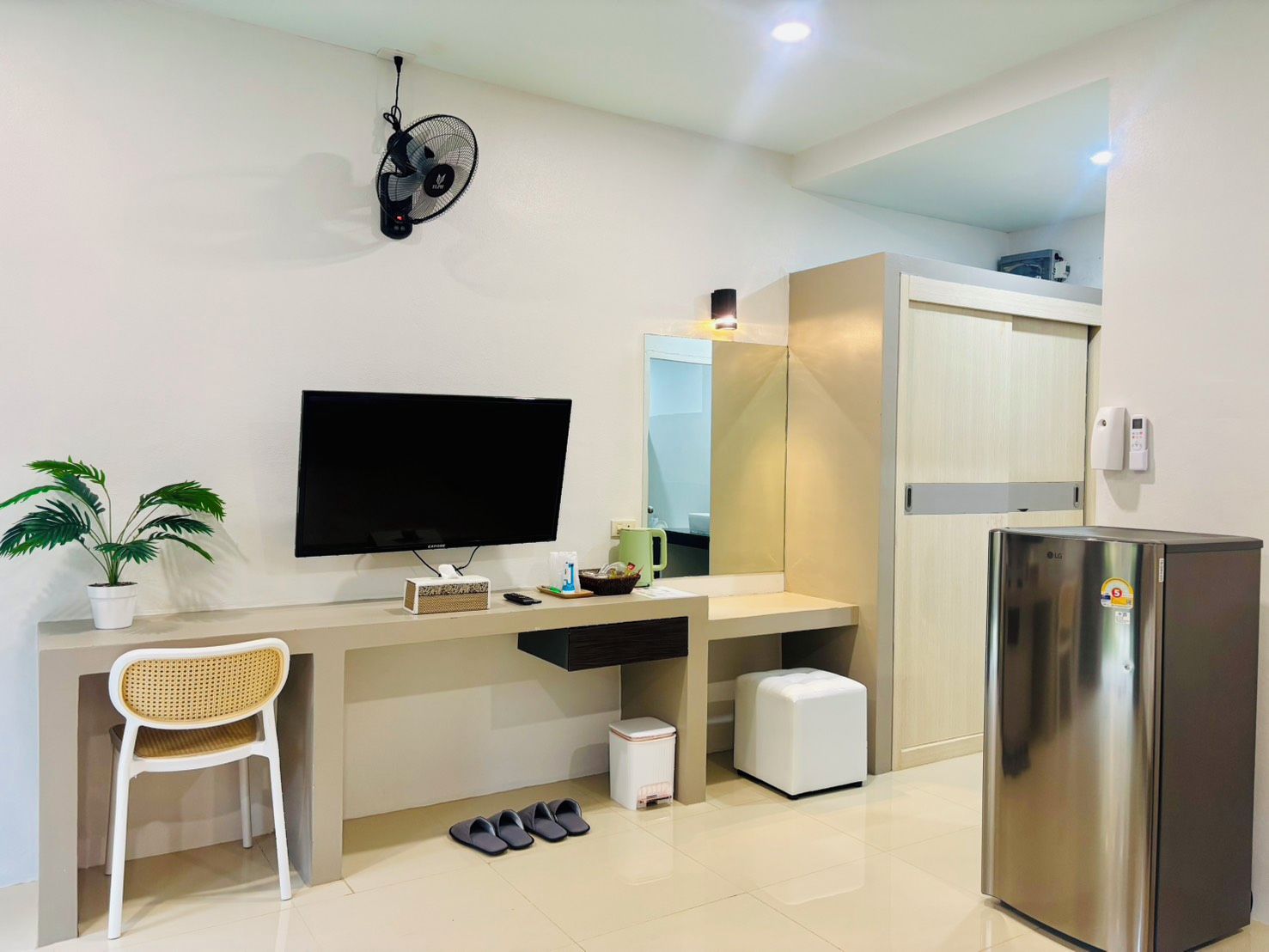 A modern room with a flat-screen TV, a small desk, a chair, a potted plant, a fan, a mirror, a refrigerator, and a trash bin.