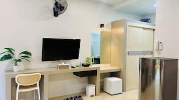 A modern room with a flat-screen TV, a small desk, a chair, a potted plant, a fan, a mirror, a refrigerator, and a trash bin.