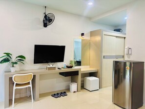 A modern room with a flat-screen TV, a small desk, a chair, a potted plant, a fan, a mirror, a refrigerator, and a trash bin.