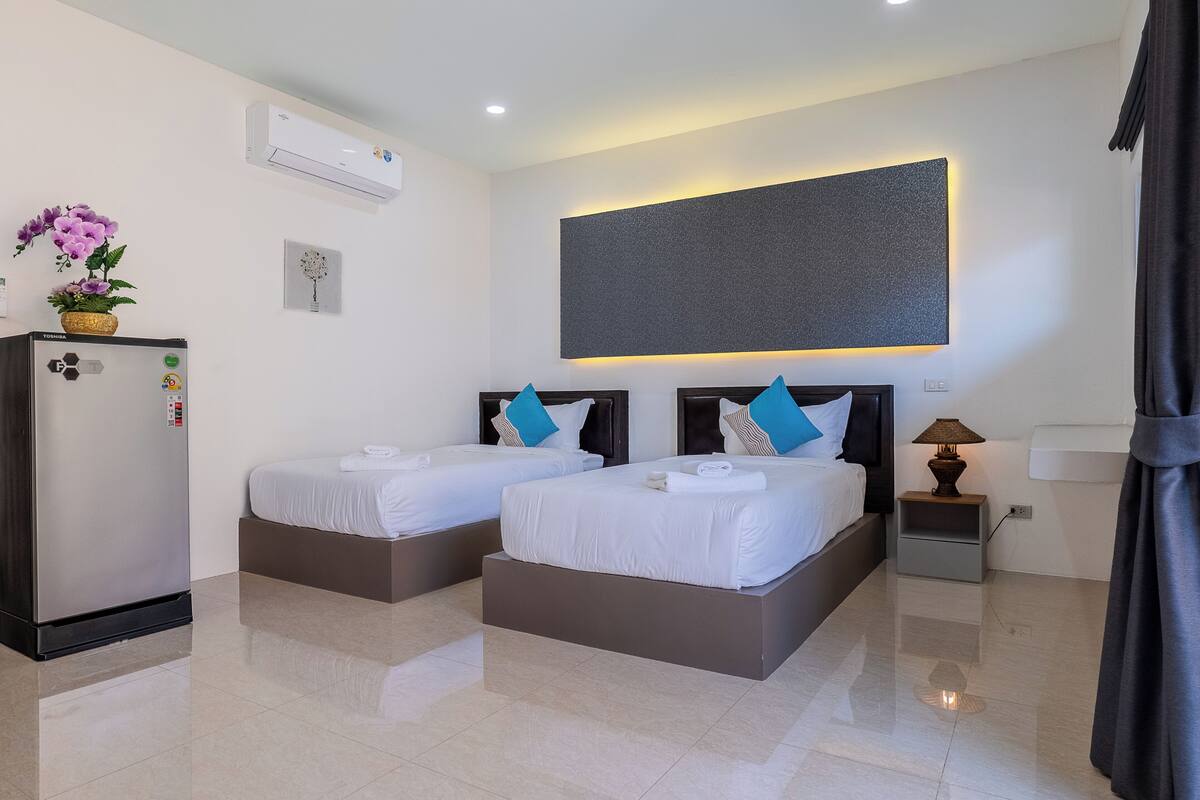 A hotel room with two beds, a refrigerator, a wall-mounted air conditioner, a bedside lamp, and a large abstract painting.