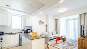 A modern kitchen with a built-in island, a dining area with a sofa and coffee table, and a living room with a TV and large windows. - Superhost  Stylish 1BR in Bloom Tower (Dubai)