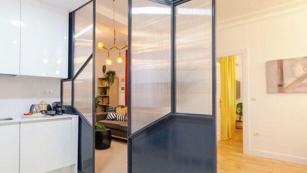 A modern kitchen with white cabinets, a black-framed glass partition, and a dining area with a sofa and cushions. - Palacio Premium Stay (Madrid)