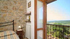 A bedroom with a stone wall, a wooden bed, a bedside table with a lamp, a radiator, and a view of a green landscape through a balcony. - Room 'Camera 3 - Campo Ruffaldo' with Shared Pool, Wi-Fi and Air Conditioning (Massa Marittima)