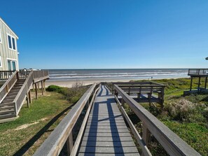 Eine Holzpromenade führt zu einem Strand, links davon befindet sich ein Gebäude.