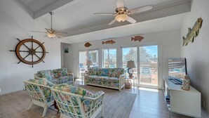 A spacious living room with a large window, a ceiling fan, a sail wheel decoration, and a view of the beach. - Pelican Place close to beach (Port Aransas)