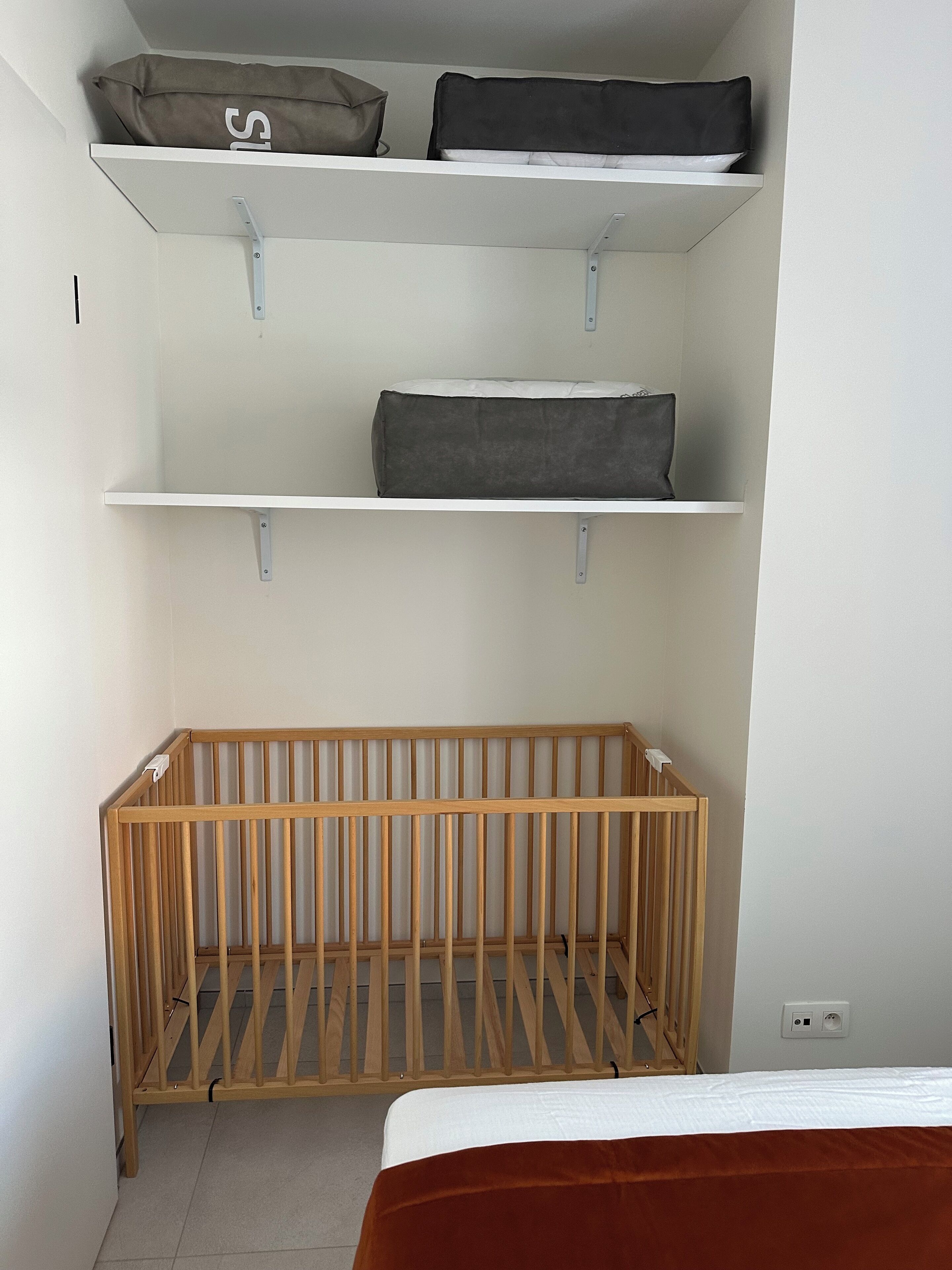 Quarto com berço de madeira, prateleiras brancas e cama com cobertor marrom.