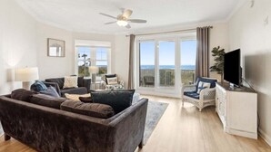 A living room with a sofa, a television, a ceiling fan, and a view through large windows. - Ocean Place 7 ~ Shore to Please (Fernandina Beach)