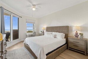 A bedroom with a large bed, a ceiling fan, a nightstand with lamps, a desk, and a view of the ocean through a sliding door.