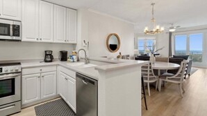 A modern kitchen with white cabinets, a built-in microwave, a coffee maker, and a dishwasher. There is a dining area with a round table and chairs, and a large window offering a view. - Ocean Place 7 ~ Shore to Please (Fernandina Beach)