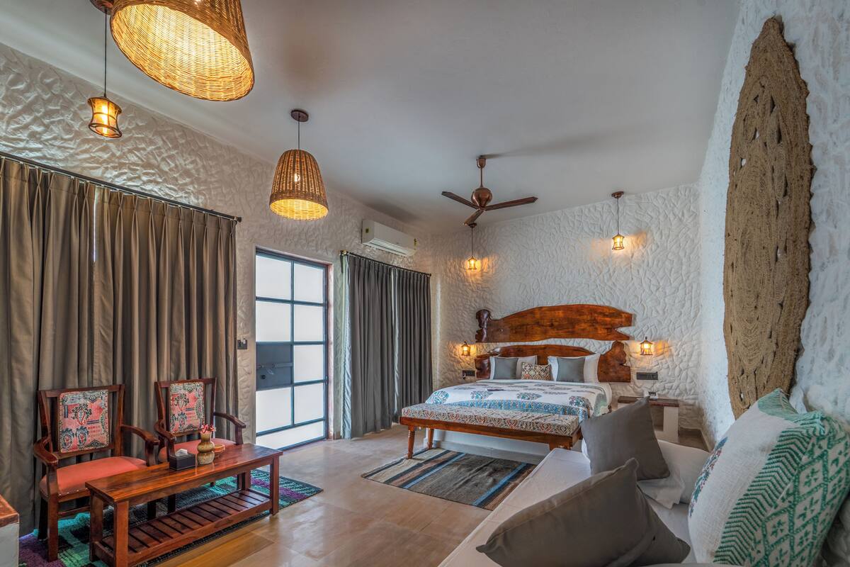 A spacious bedroom with a large bed, a wooden bench, and two chairs. There are two hanging lights and a ceiling fan.