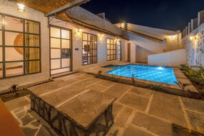 A stone pool with a thatched roof, stone walls, and a staircase leading up to a white building with large windows and wall-mounted lights. - Dunes and You, Pushkar - A Luxury Pool Villas Retreat (Pushkar)