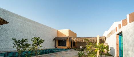 A courtyard with a swimming pool, surrounded by white walls and a thatched-roof structure.