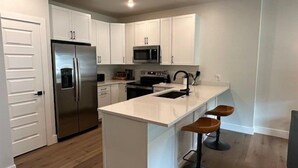 A modern kitchen with white cabinets, a stainless steel refrigerator, a microwave, and a sink. There are two stools at the island. - Premier 1BR + Den w/ Pool & Hot Tub Access (Keystone)