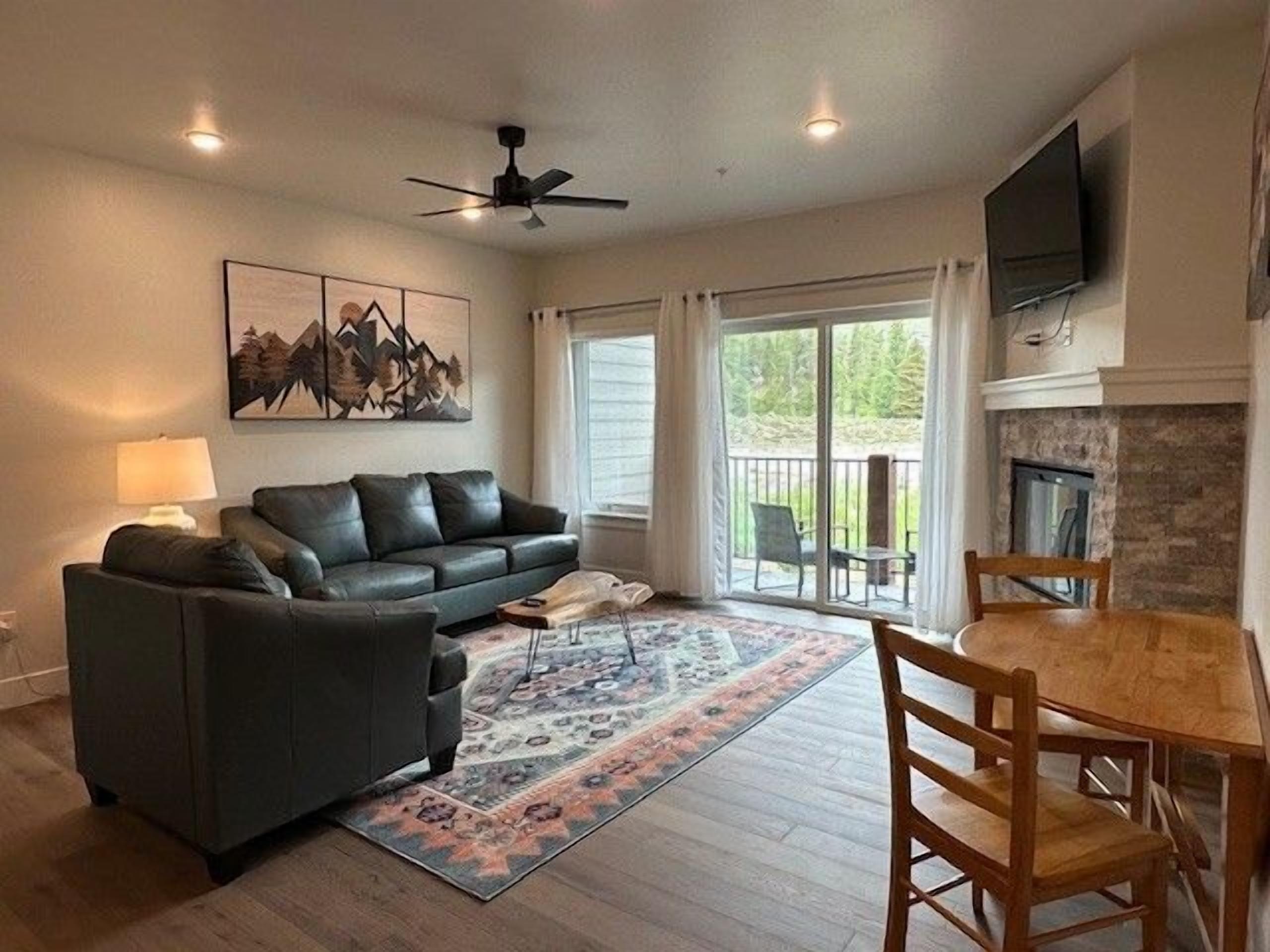 A living room with a black leather sofa, a coffee table, and a rug. There is a fireplace, a TV mounted on the wall, and a ceiling fan.
