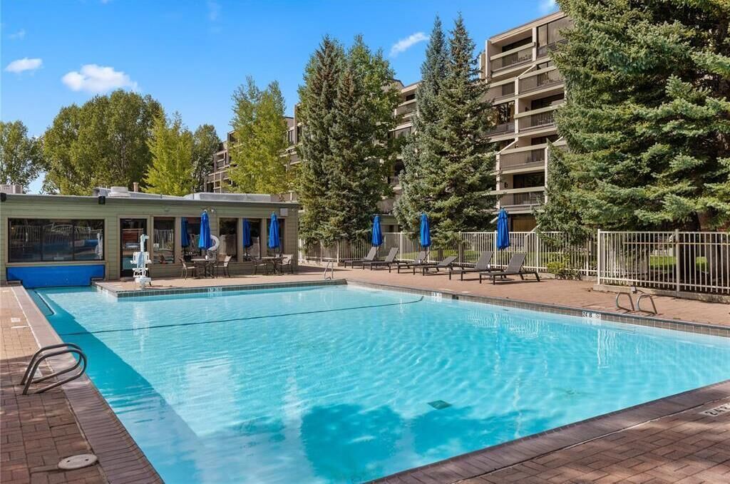 Una piscina rettangolare con superficie d'acqua blu trasparente, circondata da lettini e ombrelloni, sullo sfondo un edificio con balconi e alberi.