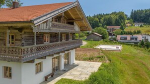 A two-story wooden house with a balcony, a stone pathway, and a view of a green hillside with other houses. - Holiday Home 'Englburger Hof' with Private Terrace, Private Garden and Wi-Fi (Tittling)
