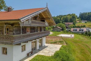 Ein zweistöckiges Holzhaus mit einem Balkon, einem Steinweg und Blick auf einen grünen Hügel mit weiteren Häusern.