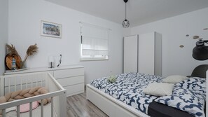 A bedroom with a bed, a crib, a window with blinds, a radiator, a wall-mounted lamp, and a framed picture.