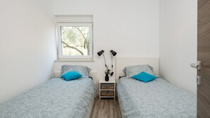 A small, well-lit room with two beds, a window, and a nightstand with lamps.
