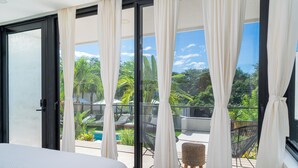 Balcón con vista a una piscina y follaje, una silla de mimbre y una cama con cabecera blanca.