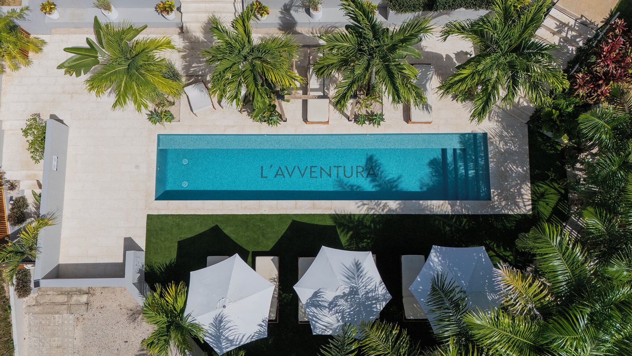 Vista aérea de una piscina señalizada como 'L'AVENTURA', rodeada de palmeras y sombrillas.