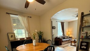 Zona pranzo con tavolo rotondo in legno e sedie, una ventola a soffitto e vista su un soggiorno attraverso un'apertura.