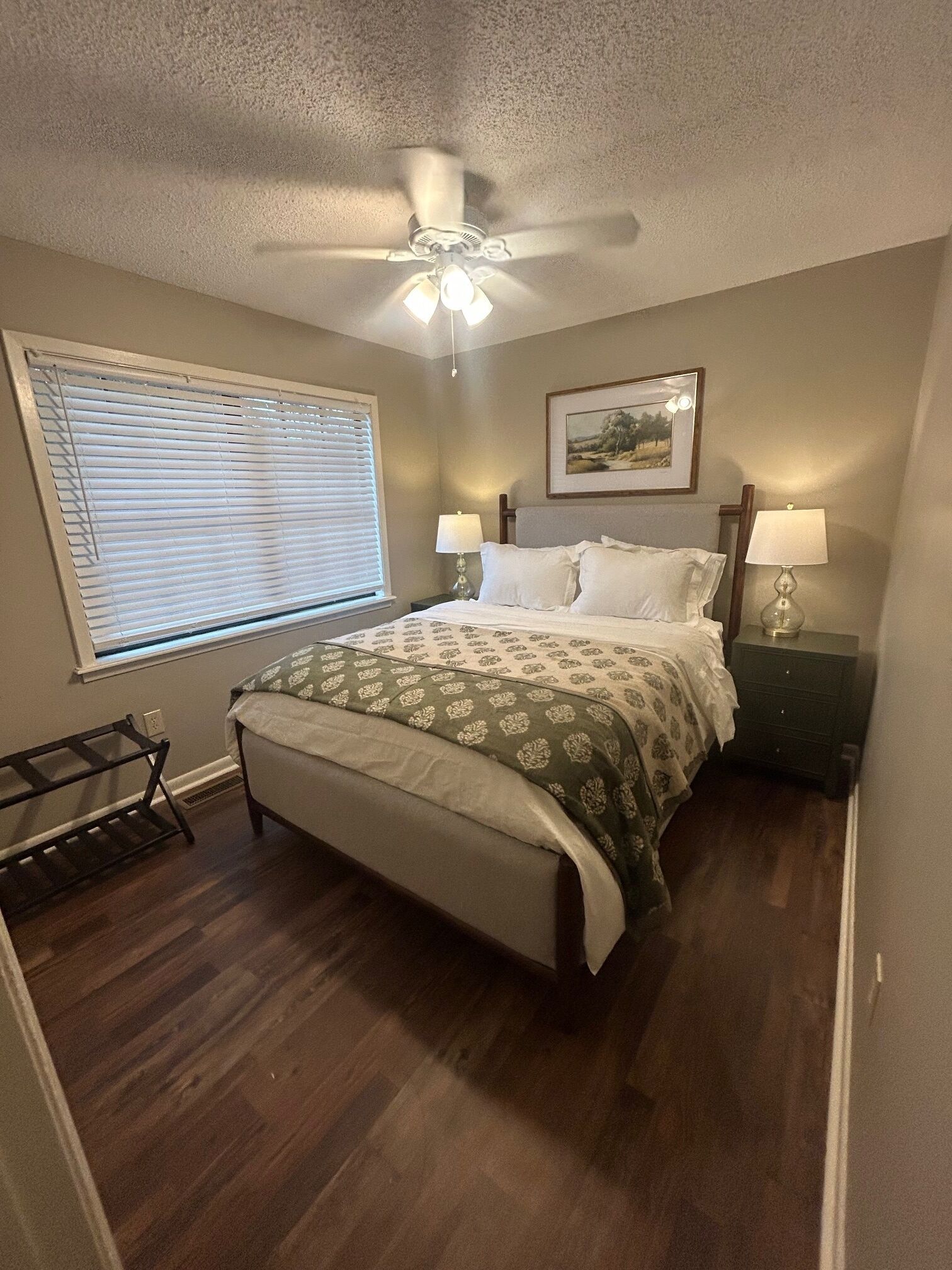 A bedroom with a bed, a ceiling fan, a window with blinds, a bedside table with lamps, and a framed picture on the wall.
