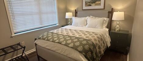 A bedroom with a bed, a ceiling fan, a window with blinds, a bedside table with lamps, and a framed picture on the wall.