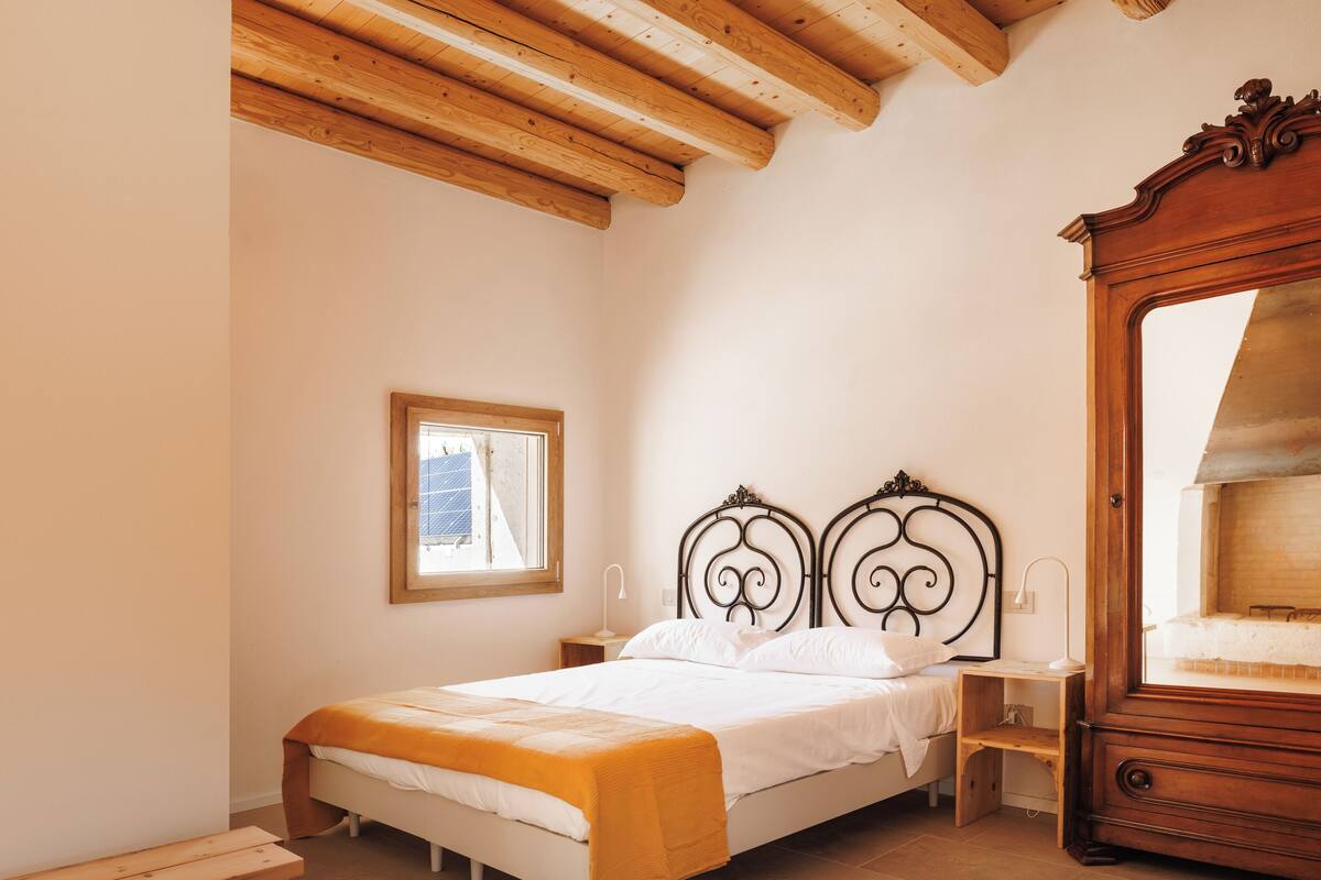 A bedroom with a wooden ceiling, a bed with white bedding, a bedside table, a nightstand, a wooden dresser, and a window.