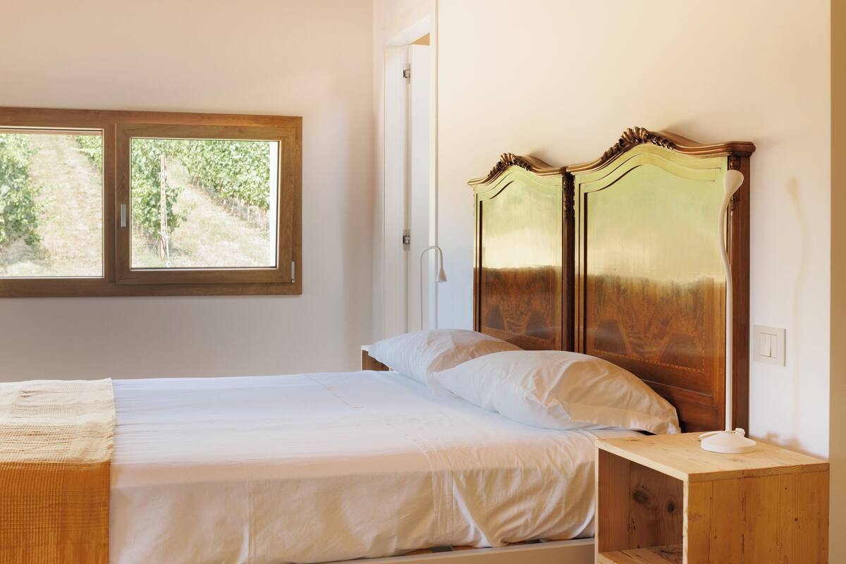 A bedroom with a wooden ceiling, a bed with white bedding, a nightstand, and a window with a view of greenery.