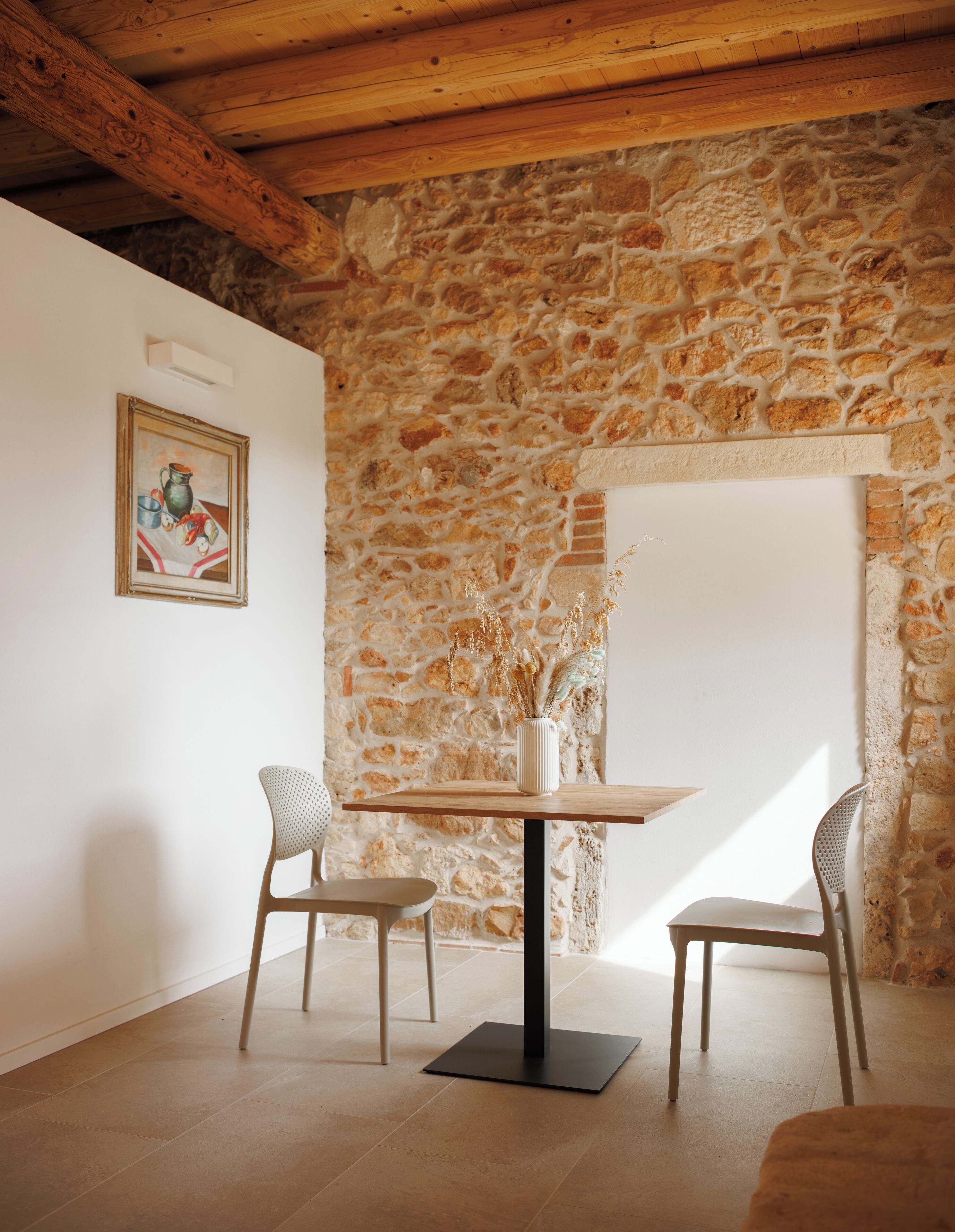 A room with a stone wall, a wooden ceiling, a table with chairs, and a framed picture on the wall.