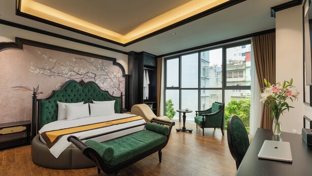 Una habitación de hotel moderna con una cama grande, un sofá verde, un escritorio con una computadora portátil y vistas de la ciudad a través de amplios ventanales.