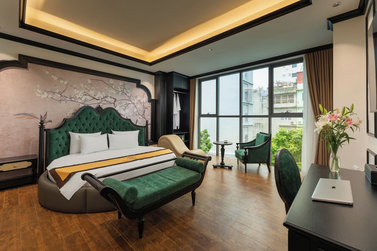A modern hotel room with a large bed, a green sofa, a desk with a laptop, and a view of the city through large windows.