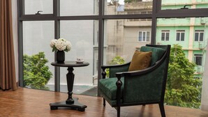 Una habitación con un sillón verde, una mesita redonda con un jarrón de flores y un ventanal que ofrece vistas a edificios y árboles.