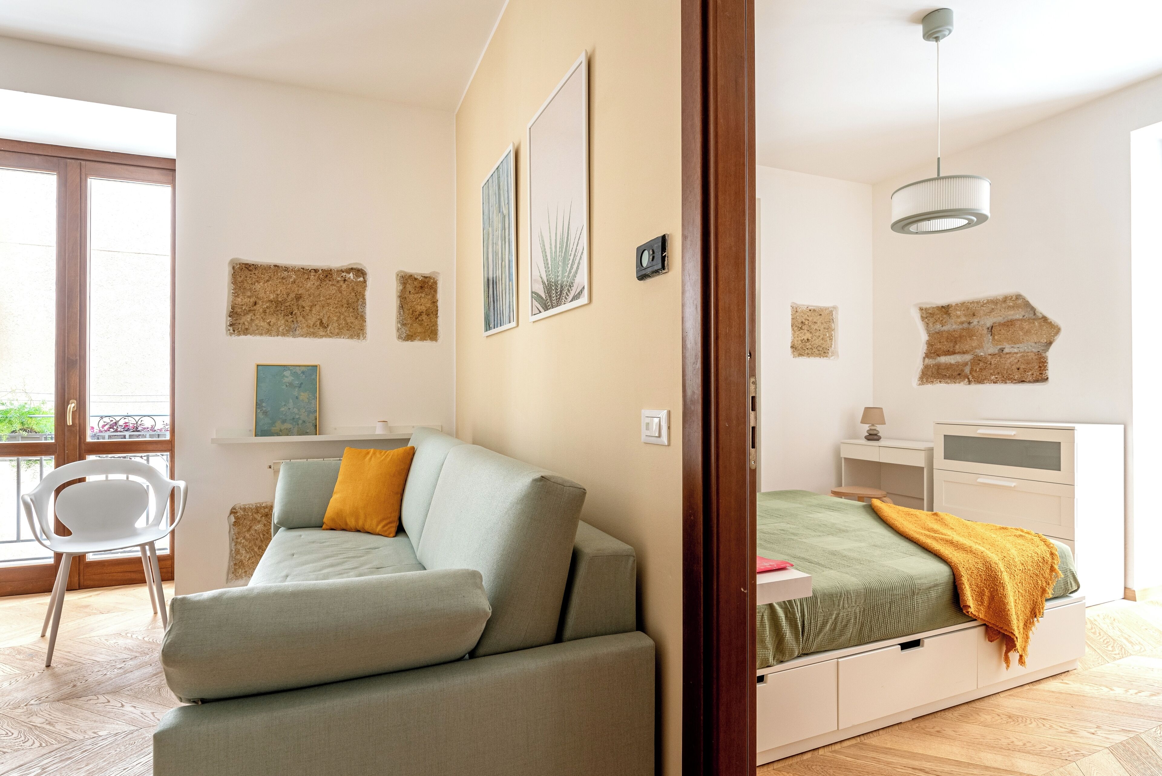 A bedroom with a sofa, a bed with a green bedspread, a white bedside table, and a wall-mounted light fixture.