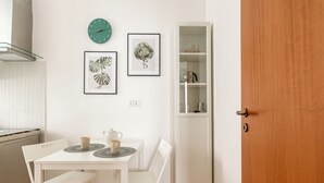 Ein kleiner, weißer Essbereich mit Tisch und Stühlen, einer Uhr, gerahmten Pflanzenbildern und einem weißen Schrank mit Regalen.