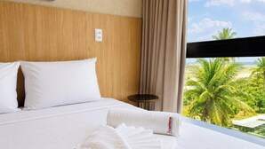 Ein Hotelzimmer mit Bett, kleinem Tisch, Fenster mit Blick auf Palmen und Strand.