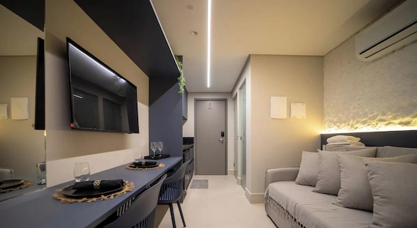 Quarto de hotel moderno com televisão de tela plana, área de refeições com mesa e cadeiras, um sofá e um aparelho de ar condicionado.