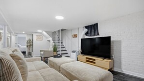 Een moderne woonkamer met een grote flatscreen-televisie, beige gestoffeerde banken, een trap en een witte baksteenmuur.