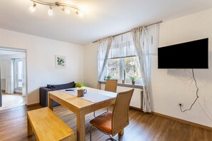 A dining area with a wooden table, chairs, a bench, a flat-screen TV mounted on the wall, and a window with curtains.