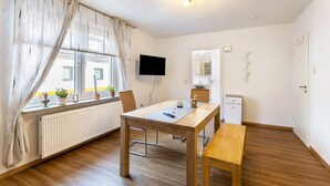 Una habitación con una mesa de madera, un banco, un televisor, un radiador, una ventana con cortinas y una puerta.