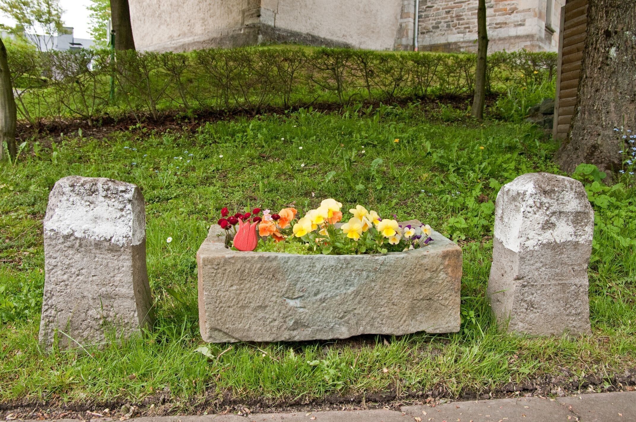 A stone planter with flowers in a grassy area, surrounded by two stone markers.