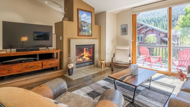 A modern living room with a fireplace, a television, and a large window overlooking a forested area. - Bright Spacious Condo with Spa Access 638 (Mammoth lakes)