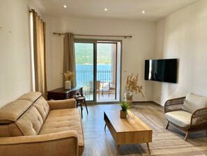 A living room with a sofa, a wooden coffee table, and a wicker chair. There is a TV mounted on the wall and a sliding glass door leading to a balcony with a view of the sea. - Cas'Antica (Propriano)