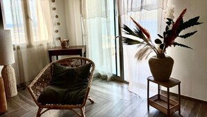 Huoneessa on rottinkituoli, pieni pöytä, lamppu ja ruukussa oleva kasvi, ja ikkunan edessä on läpikuultavat verhot.