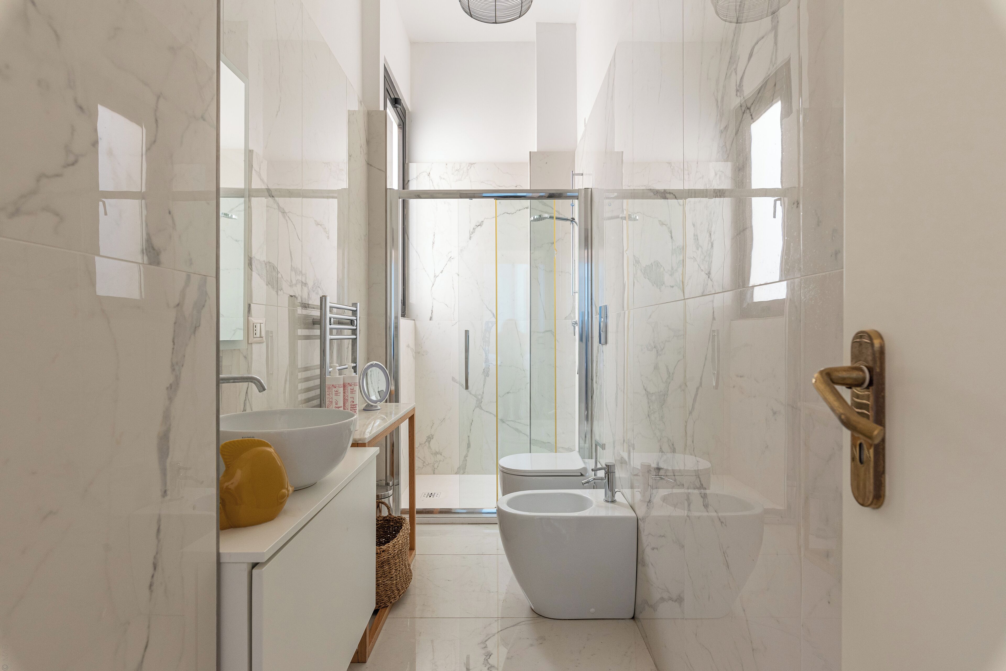 A modern bathroom with a marble floor, a white sink, a bidet, and a glass shower enclosure.
