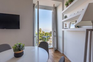 Une chambre moderne dotée d’un téléviseur à écran plat fixé au mur, d’une table à manger avec des chaises, d’une plante en pot et d’une vue sur un balcon aménagé avec du mobilier d’extérieur.